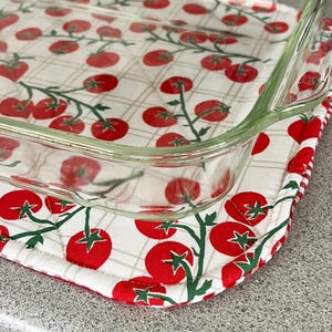May include: A clear glass baking dish rests on a quilted pot holder. The pot holder features a white background with a red tomato pattern and green stems. The edge of the pot holder is red and white striped.