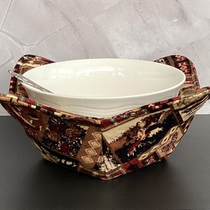 May include: A white ceramic bowl with a spoon rests inside a patterned fabric bowl cozy. The cozy features a design with brown, red, and tan colors. The cozy is designed to hold the bowl and keep the contents warm.