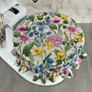 May include: A floral patterned mixer cover featuring pink, yellow, and blue flowers with green leaves and butterflies. The cover has a ruffled edge and is designed to fit over a stand mixer.