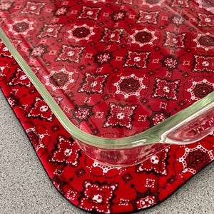 May include: A clear glass baking dish rests on a red, patterned fabric hot pad. The fabric features a repeating paisley-like design in white, black, and red. The hot pad is designed to protect surfaces from heat.