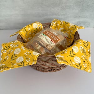 May include: A woven wicker basket holds a package of Classic Italian Table Bread. The basket is lined with four yellow fabric napkins with a white floral pattern. The napkins are folded over the basket's edges.