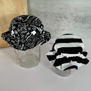 May include: Two glass jars with fabric jar covers. One cover is black with white text and illustrations, including the words "Bake with Love." The other cover is black and white striped. Both covers have ruffled edges.