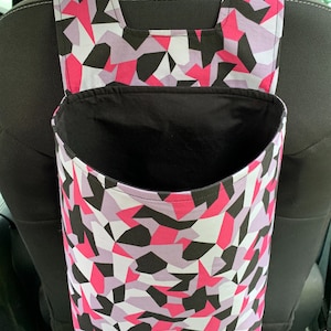 May include: A pink, black, and white geometric patterned car trash bag hangs from the headrest of a car seat. The bag has a black lining and a wide opening.