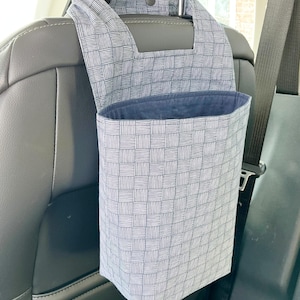 May include: A car seat organizer with a blue and white checkered pattern. The organizer hangs from the headrest and has a deep pocket. The interior is a solid navy blue. A seatbelt is visible in the background.