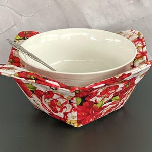 May include: A white ceramic bowl with a silver spoon rests inside a floral print bowl cozy. The cozy features a red and white floral pattern with green leaves. The bowl cozy has two handles and is designed to hold a bowl.