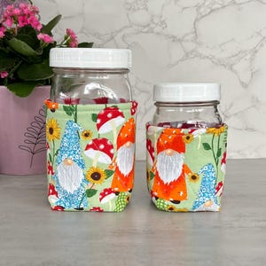 May include: Two glass jars with white lids, each wrapped in a fabric cozy. The cozies feature a whimsical pattern of gnomes, mushrooms, and sunflowers on a light green background. The jars are on a light gray surface.