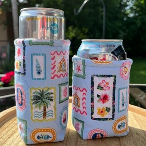 May include: Two light blue quilted can coolers with a tropical print. The print features framed images of palm trees, seashells, and flowers. One cooler holds a silver can with the text "Pur Aqua". The other holds a can with the text "Limited Edition".