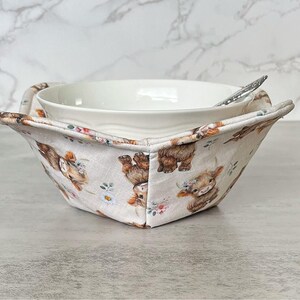 May include: A fabric bowl cozy featuring a repeating pattern of brown highland cows and small white flowers on a light beige background, holding a plain white ceramic bowl with a silver spoon resting inside the bowl.