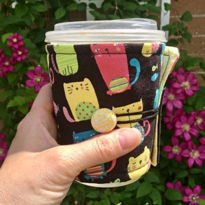 May include: A black fabric coffee cup cozy with a colorful cat print. The cozy has a yellow button and is wrapped around a clear plastic cup.