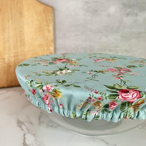 May include: A light blue bowl cover with an elastic edge, adorned with a floral pattern in pink, white, and yellow. The cover is placed over a clear glass bowl. A wooden cutting board is in the background.