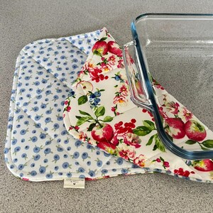 May include: A quilted fabric casserole dish carrier featuring a white background with a red apple and green leaf pattern on one side and a blue berry pattern on the other. A clear glass rectangular baking dish rests on top of the fabric carrier.