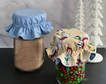 May include: Two glass jars with fabric covers. One jar contains a brown powder and is covered with a light blue fabric lid. The other jar is filled with red and green candies and has a snowman-themed fabric lid.