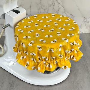 May include: A yellow mixer cover with a repeating pattern of white and black bees. The cover has a ruffled edge and is placed on top of a white stand mixer. The mixer is white.