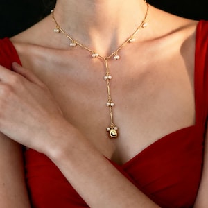 Puede incluir: Un collar dorado con pequeñas perlas redondas. El collar tiene un diseño en forma de Y con un colgante colgante. La persona de la imagen lleva un vestido rojo.