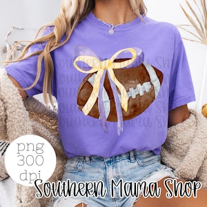 May include: A purple t-shirt with a watercolor football tied with a yellow and purple bow. The text "Southern Mama Shop" is printed on the bottom of the shirt.