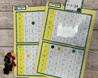 May include: Two yellow file folders with letter grids, colorful glass beads, and a label that reads "Teena Fisher" and "1-1-24." The letter grids have highlighted squares and colored magnetic dots. The folders are for educational activities.