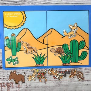May include: A blue, two-panel educational toy with a desert scene and animal cutouts. The scene includes a sun, cacti, and sand dunes. The text reads, "I can put animals in the desert." Animal cutouts include a fox, lizard, turtle, and others.