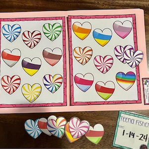 May include: A pink file folder with heart-shaped designs in various colors and patterns. Several heart-shaped cutouts are scattered around the folder. A small card with the name "Teena Fisher" and the date "1-14-24" is also present.