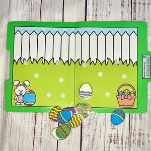 May include: A green and white folder game with a bunny holding a blue and white striped egg. The folder has a white picket fence and green grass with white polka dots. The game includes a basket with a pink bow and a blue and white striped egg. The folder has the text "Egg-cellent Learning" on the side.