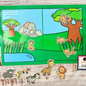 May include: A children's interactive book with a green cover and illustrations of a savanna scene. The book includes a blue pond, a tree, and various animal cutouts, including an elephant, lion, and giraffe. Text on the cover reads, "I can put animals in the grasslands."