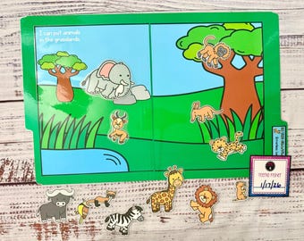 Może przedstawiać: Interaktywna książka dla dzieci z zieloną okładką i ilustracjami sceny z sawanny. Książka zawiera niebieski staw, drzewo i różne wycinanki zwierząt, w tym słonia, lwa i żyrafę. Tekst na okładce brzmi: „Mogę umieszczać zwierzęta na łąkach”.