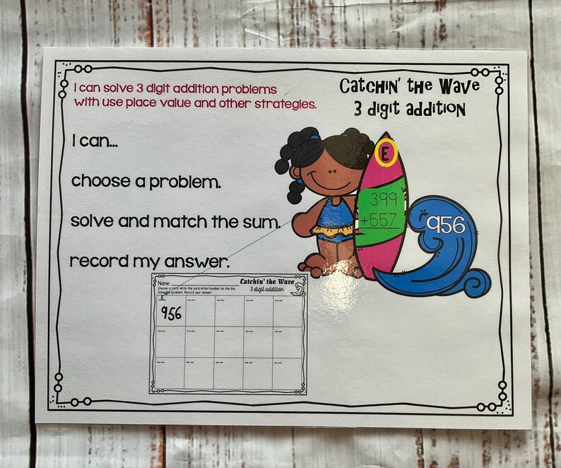 Op de afbeelding: Een zwart-wit werkblad met de titel "Catchin' the Wave 3 digit addition". Het werkblad heeft een cartoonafbeelding van een persoon op een surfplank met de woorden "Ik kan... een probleem kiezen. oplossen en de som matchen. mijn antwoord opschrijven." Het werkblad heeft een raster met het getal 956 in de linkerbovenhoek.