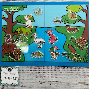 May include: A blue and green wetland-themed puzzle with various animal stickers, including a snake, frog, duck, and alligator. The text "I can put animals in the wetlands" is visible. A small card with the name "Teena Fisher" and the date "11-8-25" is in the lower left corner.