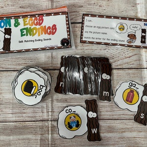 May include: A set of educational flashcards for children featuring a cartoon bacon and egg design. The cards are labeled with the words "Bacon & Eggs Endings" and include images of a boot, a cow, a gas can, and a letter 'm'. The cards are designed to help children learn about ending sounds.
