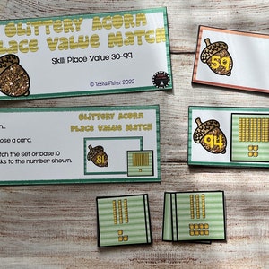 May include: A set of educational game cards featuring acorns with glitter and numbers. The cards are designed to help children learn place value. The cards include a title card that says "Glittery Acorn Place Value Match" and a set of cards with numbers and base 10 blocks.