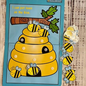 May include: A blue and yellow beehive with a branch and leaves on top. The text "I can put bees on the hive." is printed on the card. There are 9 yellow and black bees with smiling faces on the card. The text "Busy Beehive! Errorless Learning" is printed on the top right corner of the card.