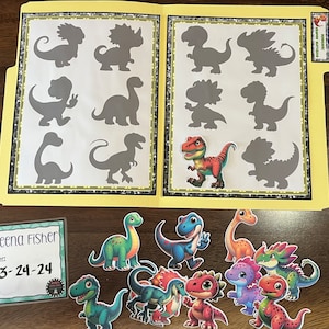 Pode incluir: Uma pasta amarela aberta para revelar duas páginas com silhuetas de dinossauros. Um adesivo de dinossauro colorido é colocado em uma página. Abaixo estão vários adesivos de dinossauros. Um rótulo diz "Teena Fisher" e "3-24-24".