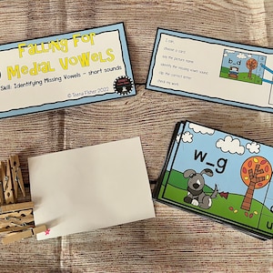 May include: A set of educational cards for children to learn about vowels. The cards feature a colorful autumn tree, a dog, and the letters a, e, i, o, u. The cards are designed to help children identify missing vowels in words. The text on the cards reads "Falling For Medial Vowels" and "Skill: Identifying Missing Vowels - short sounds".