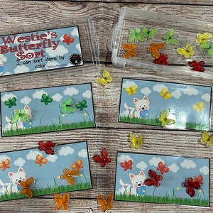 May include: A clear plastic container and four illustrated cards with a Westie dog and butterflies. The cards have a blue sky background with clouds and grass. The text on the container reads "Westie's Butterfly Sort". Assorted colorful butterfly decorations are included.