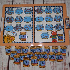 May include: A laminated file folder learning activity featuring illustrations of fish bowls with varying numbers of orange fish, a cat, and fish food containers. Includes loose cut-out fish food containers labelled with numbers 1 through 20.