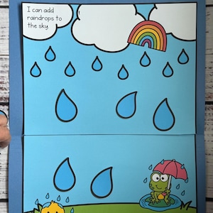May include: A blue folder with a colorful illustration of a rainy day scene. The image includes a rainbow, raindrops, clouds, a frog with an umbrella, and a duckling. Text reads "I can add raindrops to the sky" and "SPLISH SPLASH Errorless Learning".