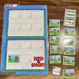May include: A blue folder with a "David and Goliath" story board. The board has numbered boxes with text and illustrations. Small illustrated cards and a name tag are scattered around the folder.