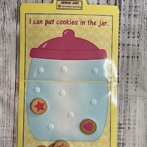May include: A yellow learning tool with a blue cookie jar illustration and the text "I can put cookies in the jar." The jar has a pink lid and white dots. Several cookie-shaped tokens with heart and star designs are included.