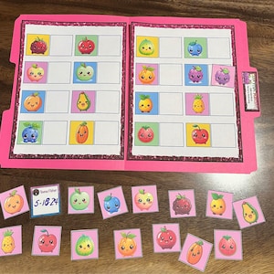 May include: A pink folder with a grid of squares, each containing a cartoon fruit illustration. Loose fruit illustrations are scattered below. The fruits include apples, oranges, berries, and a pineapple. A small label reads "Teena Fisher 5-18-24".