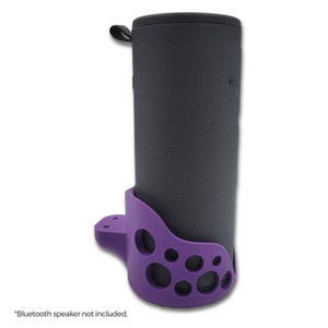 May include: Purple plastic stand designed to hold a cylindrical Bluetooth speaker. The stand has a unique design with multiple holes and a curved shape. "Bluetooth speaker not included." is written below the stand.