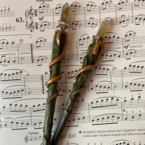 May include: Two handcrafted pens with green and gold accents, topped with a crystal. The pens are decorated with a gold-colored spiral design. The pens rest on a sheet of musical notes.
