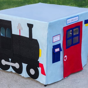 May include: A blue and white play tent shaped like a train with a red door and a blue mailbox. The tent has a black train engine on one side and the words "All Aboard" and "Meet Train 1:00 pm" on the other side.