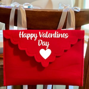 May include: Red felt envelope with white scalloped edges and the text "Happy Valentines Day" with a white heart. The envelope is hanging on a wooden chair back with white ribbon.