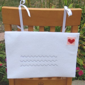 May include: White felt envelope with a red heart on the upper right corner. The envelope has a zig-zag line sewn across the front. The envelope is attached to a wooden chair with white ribbon.
