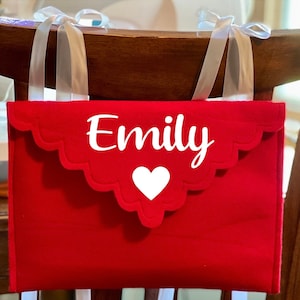 May include: Red felt chair back organizer with white scalloped edges and a white heart. The organizer has the name "Emily" written in white.