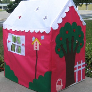 May include: A pink play tent with a white roof and scalloped edges. The tent has a window with green curtains, a birdhouse, a green tree with red apples, and a white picket fence. The tent is on a gray concrete surface.