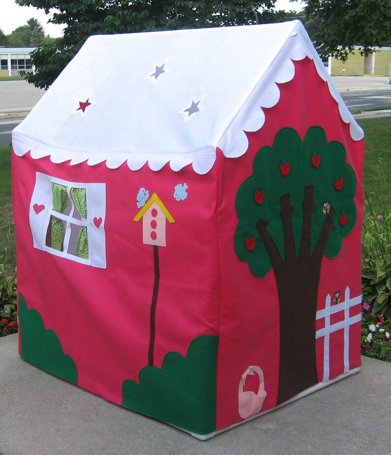 May include: A pink play tent with a white roof and scalloped edges. The tent has a window with green curtains, a birdhouse, a green tree with red apples, and a white picket fence. The tent is on a gray concrete surface.