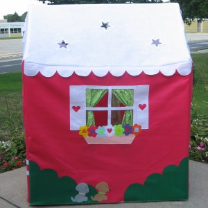 May include: A red play tent with a white roof and a window with green curtains. The tent has a scalloped edge and a flower box with colorful flowers. Two small dogs are in front of the tent.