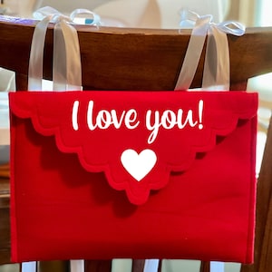 May include: A red felt envelope-shaped chair decoration with the text "I love you!" and a white heart. The top edge has a scalloped design and is attached to the chair with white ribbons.