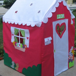 May include: A pink and white play tent with a scalloped roof, a white door with a heart-shaped window, and a green bush at the bottom. The tent has a window with green curtains and a flower box with colorful flowers. The number 1234 is on the side of the tent.