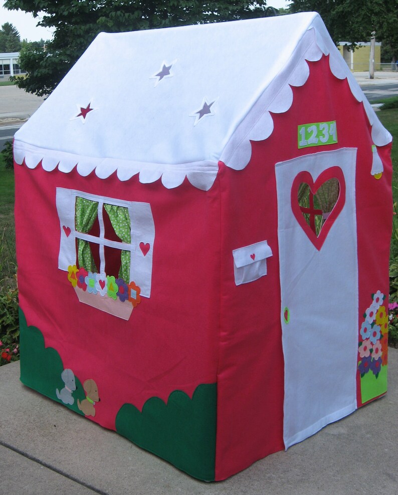 May include: A pink and white play tent with a scalloped roof, a white door with a heart-shaped window, and a green bush at the bottom. The tent has a window with green curtains and a flower box with colorful flowers. The number 1234 is on the side of the tent.
